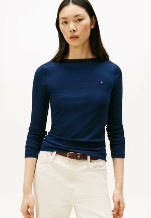 Marineblauwe geribbelde long sleeve top met een boot-formige halslijn en een klein logo op de borst, gecombineerd met lichtgekleurde jeans en een bruine riem.
