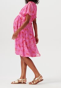 Robe de maternité rose vif avec imprimé floral, manches courtes flottantes et taille cintrée à nouer. Associée avec des sandales plates dorées.