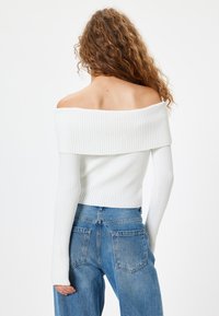 Weißer, gerippter Off-Shoulder-Pullover mit langen Ärmeln, kombiniert mit hochgeschnittenen blauen Jeans. Texturiertes Material und figurbetonter Schnitt.