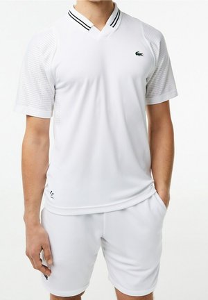 Man draagt een witte sportshirt met korte mouwen en geperforeerde mouwen, gecombineerd met witte korte broek, staand met één hand in de zak.