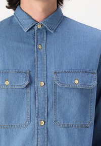 Lichtblauwe denim overhemd met een gestructureerde kraag, knoopsluiting en twee borstzakken met lichtbruine knopen en zichtbare stiksels.