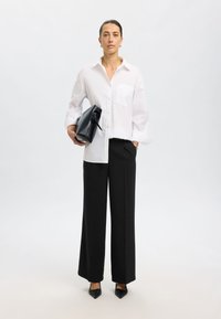 Witte blouse met knopen en een borstzak aan de linkerkant, gecombineerd met zwarte wijde broek en puntige zwarte hakken, terwijl ze een zwarte handtas vasthoudt.