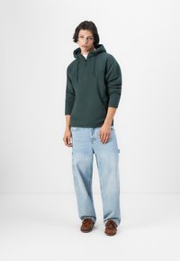 Zöld polár kapucnis pulóver elülső zsebbel, világoskék, oversized farmernadrággal és barna csónakcipővel párosítva.