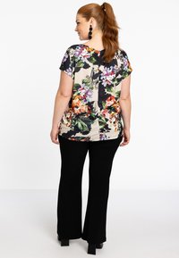 Bluse mit floralem Muster und kurzen Ärmeln in Beige, verziert mit bunten Blumen. Kombiniert mit schwarzen Schlaghosen und klobigen schwarzen Ohrringen.