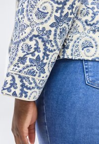 Αριστερό χέρι που φοράει λευκό μπουφάν με μπλε φλοράλ σχέδιο παϊέτας πάνω από μπλε τζιν τζιν, με το χέρι χαλαρό δίπλα.