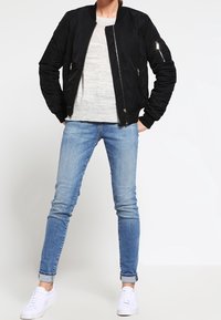 Czarna pikowana kurtka bomberka z zamykanymi na zamek kieszeniami, noszona na jasnoszarym swetrze, w połączeniu z jasnoniebieskimi jeansami typu skinny i białymi trampkami.