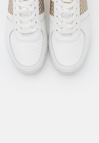 Ett par vita sportskor med vita snören och guldmönstrade detaljer på sidorna, sedd ovanifrån mot en vit bakgrund.