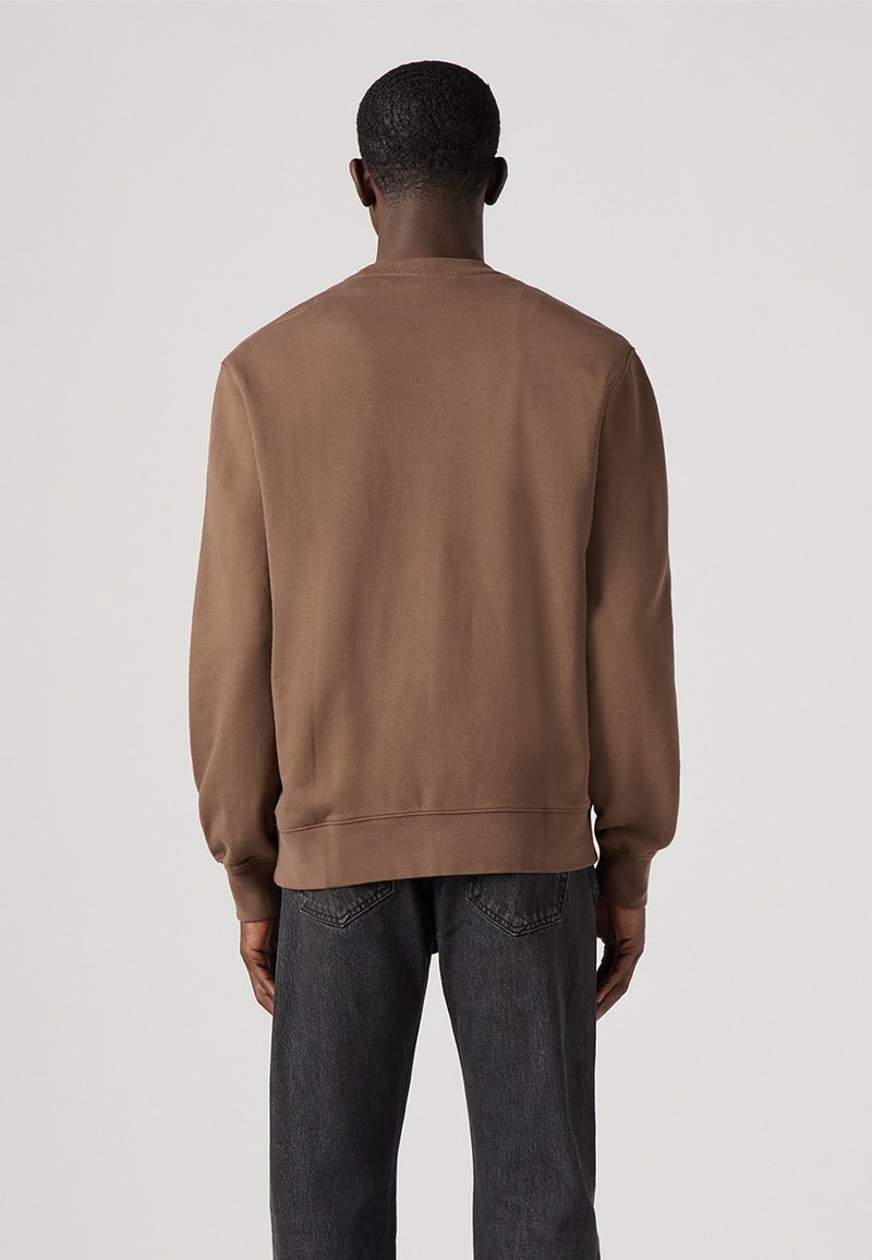 Braunfarbener Rundhalsausschnitt-Pullover aus weichem Stoff, mit gerippten Bündchen und Saum, in lässiger Passform, getragen zu dunklen Jeans.