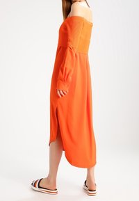 Femme en robe midi orange à manches longues et épaules dénudées avec une fente latérale, portant des sandales plates rayées, debout devant un fond blanc.