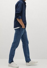 Granatowa koszula na guziki z podwiniętymi rękawami, zestawiona z niebieskimi dżinsami i białymi trampkami. Dopasowany krój; prosty design, brak widocznych wzorów czy ozdób.