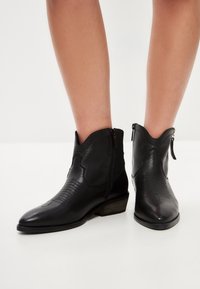 Botines de cuero negro con punta puntiaguda y borde superior con diseño festoneado. Presentan costuras texturizadas y una cremallera lateral. Tacón bajo apilado.