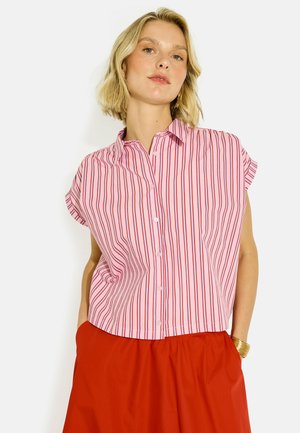 Mujer con camisa de botones de manga corta a rayas verticales rosas y rojas y pantalones rojos lisos con las manos en los bolsillos.