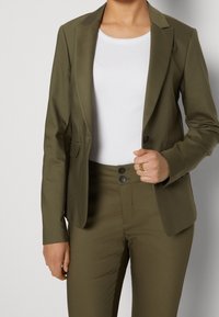 Marynarka w kolorze zielonym oliwkowym, dopasowana, z długimi rękawami i klapami z wycięciem, w zestawie z dopasowanymi spodniami; modelka ma na sobie białą obcisłą koszulę pod spodem.