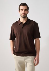 Polo shirt marron avec un col, des manches courtes et une poche sur la poitrine gauche. Fabriqué en tissu doux avec une texture lisse. Détail de patte de boutonnage à deux boutons.