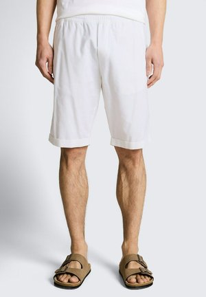 Mężczyzna ubrany w białe spodenki do kolan i brązowe sandały z klamrą, stojący na tle jasnoszarego tła.