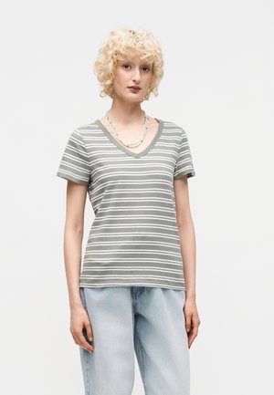 Ξανθός άνθρωπος με σγουρά μαλλιά που φορά γκρι και λευκό ριγέ T-shirt με V-λαιμόκοψη και ανοιχτούς μπλε χαλαρούς τζιν, στέκεται μπροστά σε απλό φόντο.