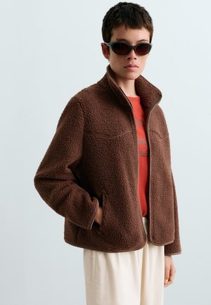 Persona che indossa occhiali da sole scuri, giacca sherpa marrone con cerniera sopra una camicia rossa e pantaloni chiari con una mano nella tasca della giacca.
