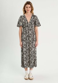 Robe portefeuille à motifs floraux, noir et crème, avec des manches courtes évasées, décolleté en V profond, longueur cheville, associée à des chaussettes blanches et des baskets beige.