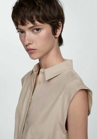Camicia beige senza maniche con colletto e una texture leggera e liscia, caratterizzata da una sottile lucentezza. I bottoni sono piccoli e di colore neutro.