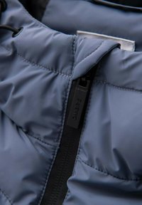 Niebieska pikowana tkanina kurtki z widocznym przeszyciem i czarnym zamkiem błyskawicznym. Na suwaku znajduje się metka z napisem "OMBRE". Gładka faktura.