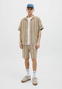 Beige kockás, rövid ujjú ing és az ahhoz illő rövidnadrág, texturált anyagból. Kiegészítve egy kék sapkával és krémszínű, barna részletekkel ellátott sportcipővel.