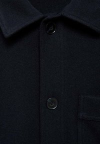 Camicia button-up in lana scura con colletto, tasca singola e un bottone nero lucido, realizzata in un tessuto morbido e testurizzato.