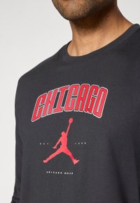 Schwarzes Langarm-T-Shirt mit rotem "CHICAGO"-Text und einer Silhouette eines springenden Basketballspielers. Glatte Stoffqualität mit Rundhalsausschnitt.