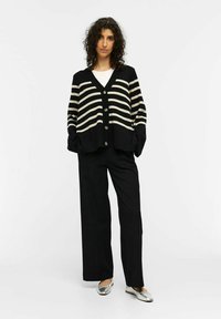 Cardigan a righe bianche e nere con scollo a V, frontale con bottoni e vestibilità ampia, abbinato a pantaloni neri a gamba larga e scarpe flat argento.
