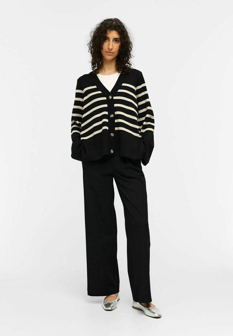 Gilet rayé noir et crème avec un col en V, devant boutonné et coupe ample, associé à un pantalon large noir et des chaussures plates argentées.