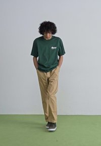 Grøn kortærmet t-shirt, beige løstsiddende bukser, grå sneakers. T-shirten har logo på brystet. Enkel, afslappet design.