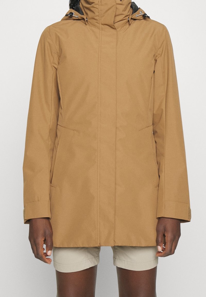 Femme portant une veste imperméable à capuche couleur sable et un short beige, debout devant un fond blanc uni.