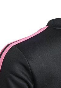 Detailní záběr na rameno černé bundy s růžovými pruhy běžícími podél švu ramene a žebrovaným límcem.