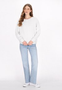 Světle šedá mikina s širokým výstřihem a spadlými rameny, doplněná o světle modré džíny. Modelka stojí na bílém pozadí.