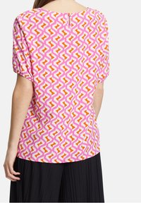Top à manches courtes rose avec un motif zigzag orange et blanc. Il présente un col rond et un détail de bouton à l'arrière.
