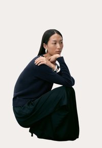 Marinblå tröja med ribbad fåll, kombinerad med en flödande svart kjol. Minimalistiska silverörhängen framhäver stilen. Neutral bakgrund.