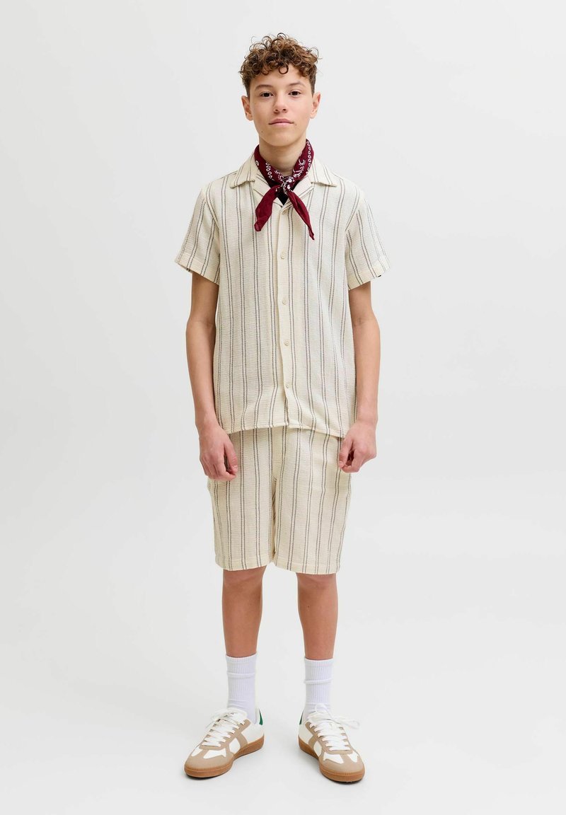 Adolescente vestido con camisa de manga corta con botones y rayas crema, pantalones cortos a juego, calcetines blancos, zapatillas deportivas y un pañuelo rojo atado alrededor del cuello.