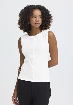 Kobieta o kręconych włosach, ubrana w białą, bezrękawnikową bluzkę na guziki i czarne spodnie, patrząca w bok na tle jasnego, jednolitego tła.