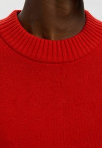 Nahaufnahme einer Person, die einen leuchtend roten, gerippten Rundhalsausschnitt-Pullover trägt, wobei der Hals und der obere Brustbereich zu sehen sind.