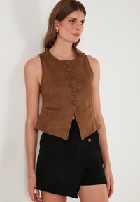 Barna nubuk ujjatlan mellény, kerek nyakkivágással, elől gombos záródással és két oldalsó zsebbel; fekete rövidnadrággal párosítva, amely oldalsó gombbal rendelkezik.