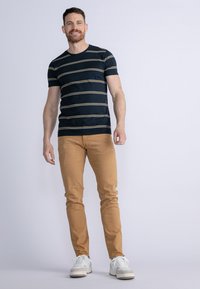 Marinrandig t-shirt med ljusgula linjer, parat med ljusbruna slim-fit byxor och vita sneakers, stående mot en enfärgad bakgrund.