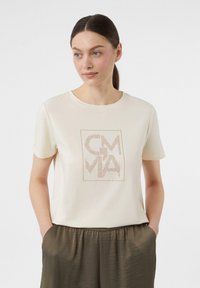 Cream T-shirt with short sleeves, featuring a square design and dotted lettering in brown. Soft fabric, relaxed fit, paired with olive green pants.