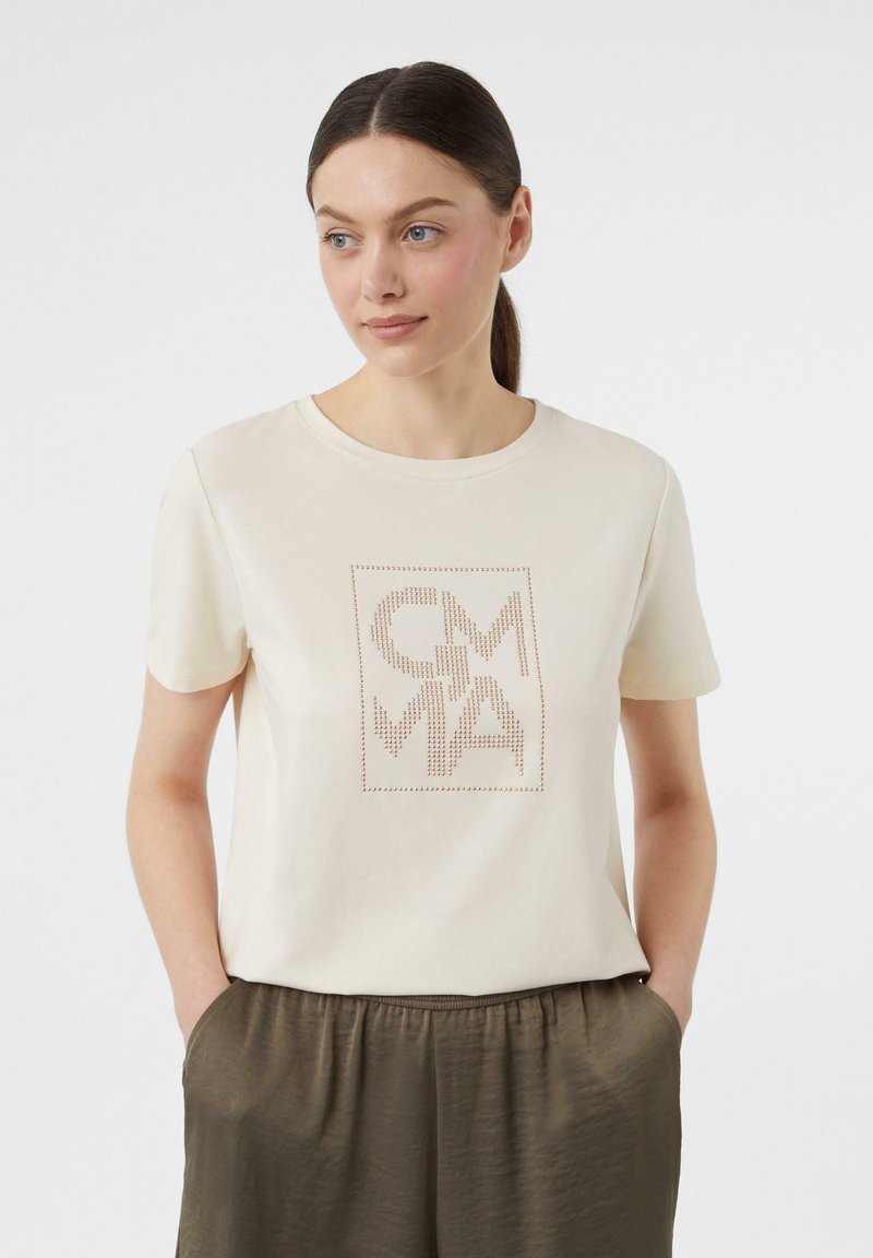 Cream T-shirt with short sleeves, featuring a square design and dotted lettering in brown. Soft fabric, relaxed fit, paired with olive green pants.