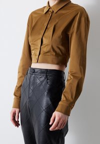 Persona che indossa una camicia a bottone corta marrone abbinata a pantaloni in pelle trapuntata nera a vita alta, su uno sfondo neutro.