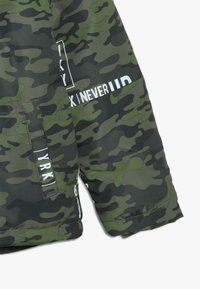 Camo-pufferjakke i forskellige nuancer af grøn og sort med stikningsdetaljer og fed hvid tekstpåføring på ærmet.