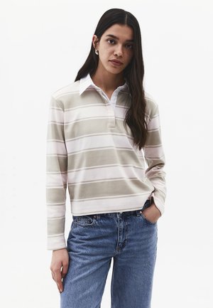 Kobieta o długich ciemnych włosach, ubrana w beżowo-białą paskowaną koszulkę polo z długim rękawem i niebieskie jeansy, stojąca z jedną ręką w kieszeni.