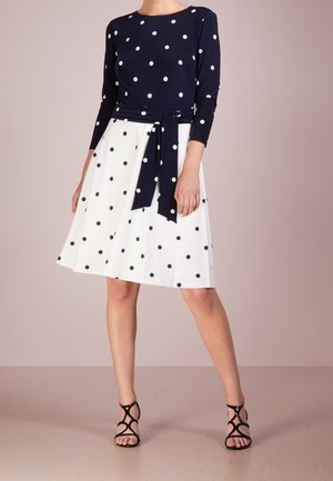 Femme portant un haut bleu marine à manches longues avec des pois blancs et une jupe blanche à pois bleu marine, associée à des talons noirs à brides.