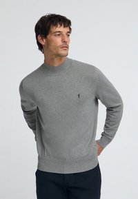 Pull gris en tricot avec un col haut, des poignets et un ourlet côtelés. Présente un petit emblème noir sur le côté gauche de la poitrine. Texture lisse partout.