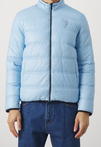 Ljusblå puffjacka med horisontell quiltning, hög krage, dragkedjestängning och en diskret logotyp. Bärs över mörk denim.