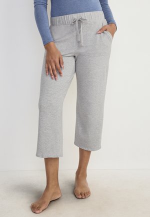 Frau trägt hellgraue, kurz geschnittene Jogginghose mit elastischem Bund und Kordelzug, kombiniert mit einem blauen Langarmshirt, barfuß.