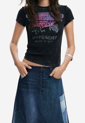 Frau trägt ein schwarzes T-Shirt mit dem Aufdruck "To be different you look so good" und einen Patchwork-Denim-Rock mit Knopfleiste vorne, Hände in den Taschen.